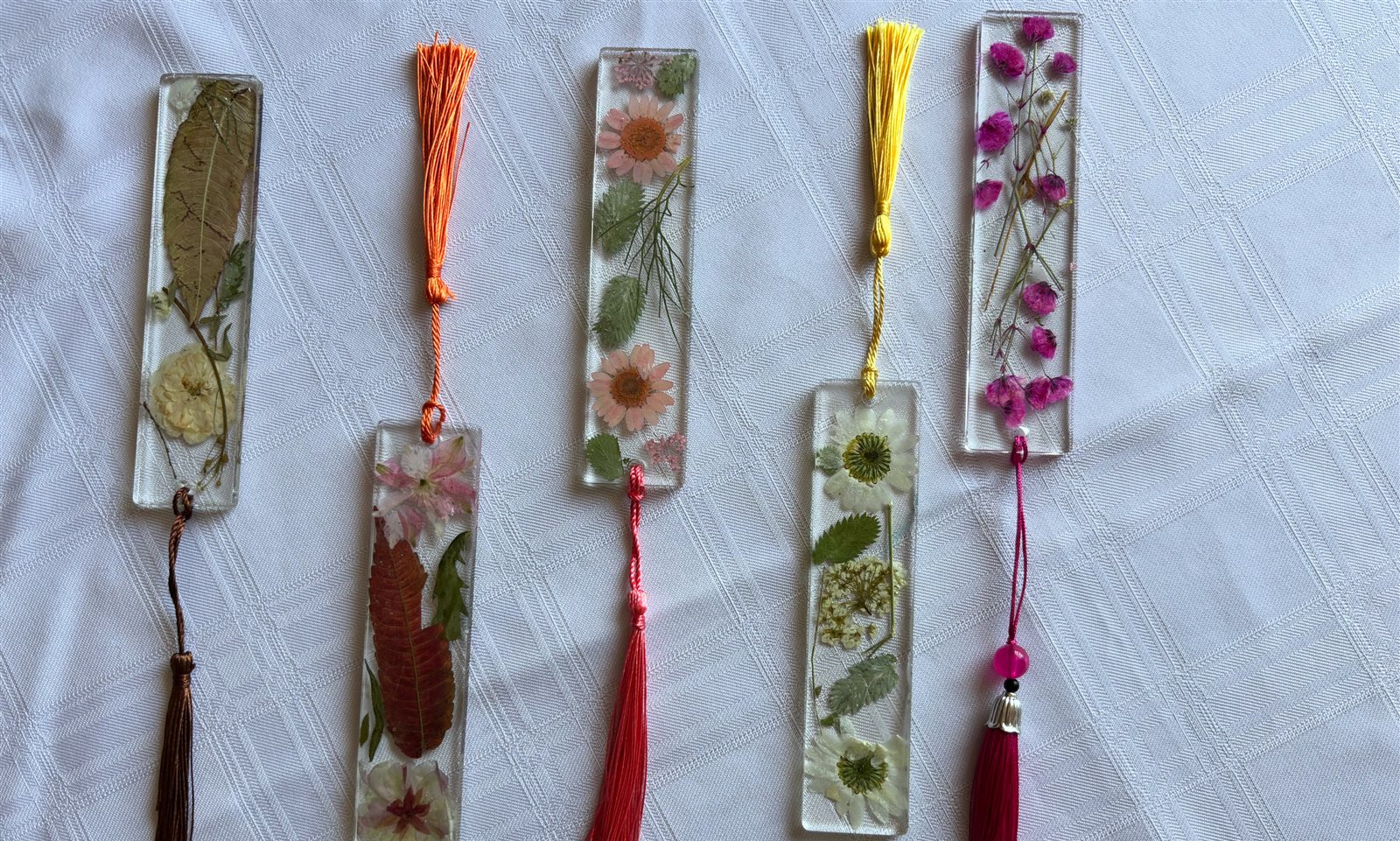 Five clear resin bookmarks holding pressed flowers and leaves, each with a coloured tassel